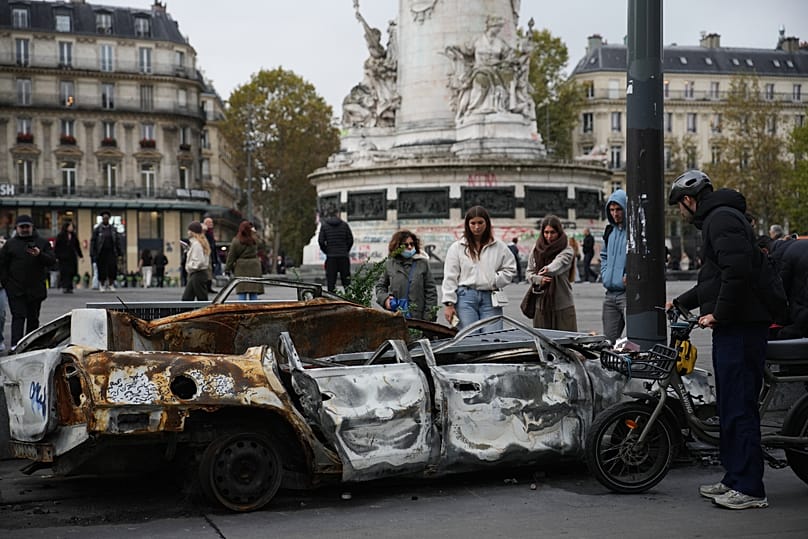 Des passants devant une carcasse de voiture calcinée, place de la République, à Paris, mardi 20 octobre 2026.