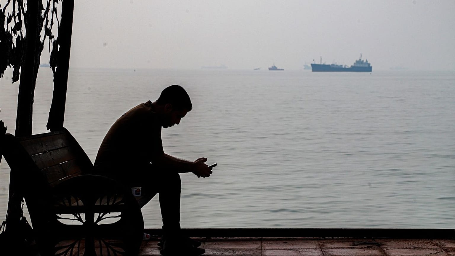 Um homem olha o telemóvel num abrigo costeiro enquanto navios comerciais surgem ancorados no estreito de Ormuz, perto de Bandar Abbas, Irão, 6 de outubro de 2026