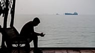 A man checks his cellphone at a waterfront shelter as commercial vessels appear anchored in the Strait of Hormuz off Bandar Abbas, Iran, Monday, Tuesday, Oct, 6, 2026. (Amirho