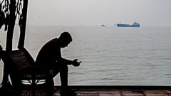 A man checks his cellphone at a waterfront shelter as commercial vessels appear anchored in the Strait of Hormuz off Bandar Abbas, Iran, Monday, Tuesday, Oct, 6, 2026. (Amirho