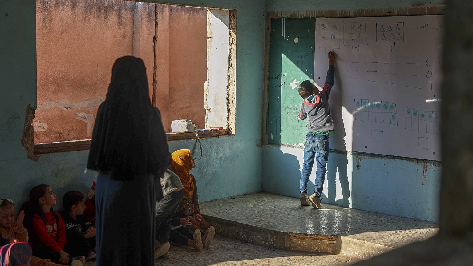 يكتب أحد التلاميذ على السبورة البيضاء بينما يراقبه زملاؤه ومعلمهم خلال درس داخل فصل دراسي في مدرسة "معر شمارين" الابتدائية، التي تحمل جدرانها آثار ثقوب خلفتها الرصاصات إبان ال