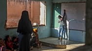 يكتب أحد التلاميذ على السبورة البيضاء بينما يراقبه زملاؤه ومعلمهم خلال درس داخل فصل دراسي في مدرسة "معر شمارين" الابتدائية، التي تحمل جدرانها آثار ثقوب خلفتها الرصاصات إبان ال